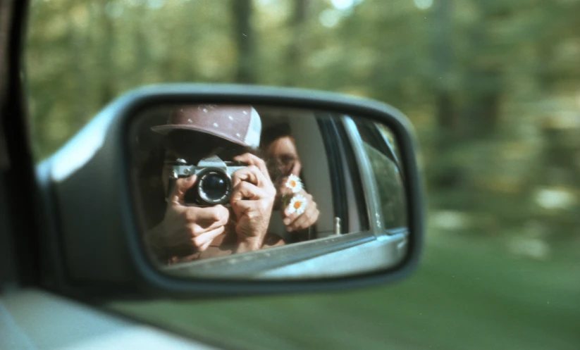 person holding camera and taking picture of the other woman through a rear view mirror