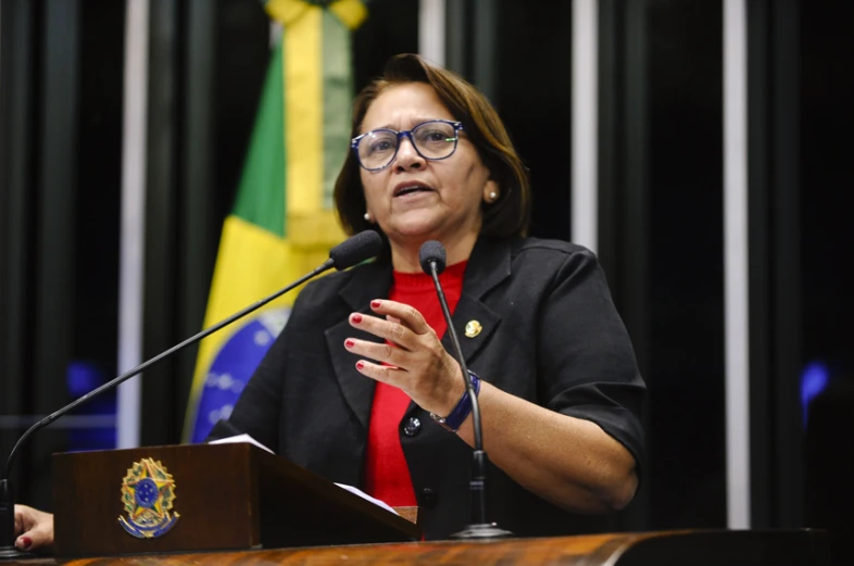 a woman in glasses sitting at a podium talking