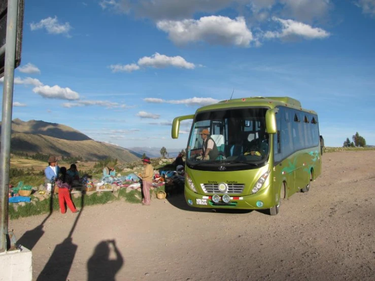 a bus sits parked on a dirt road with people standing around it