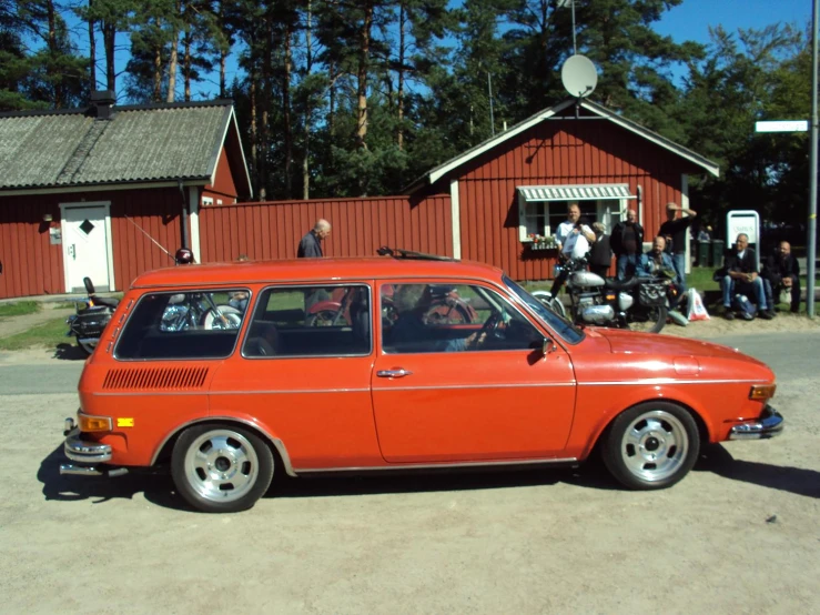 an orange station wagon is parked outside of a building with some people on bikes and a car