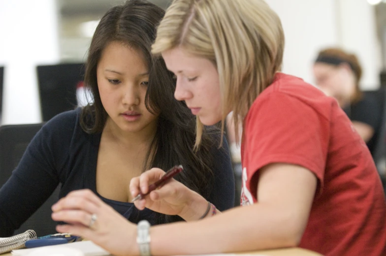 two girls looking at a paper while using a cell phone