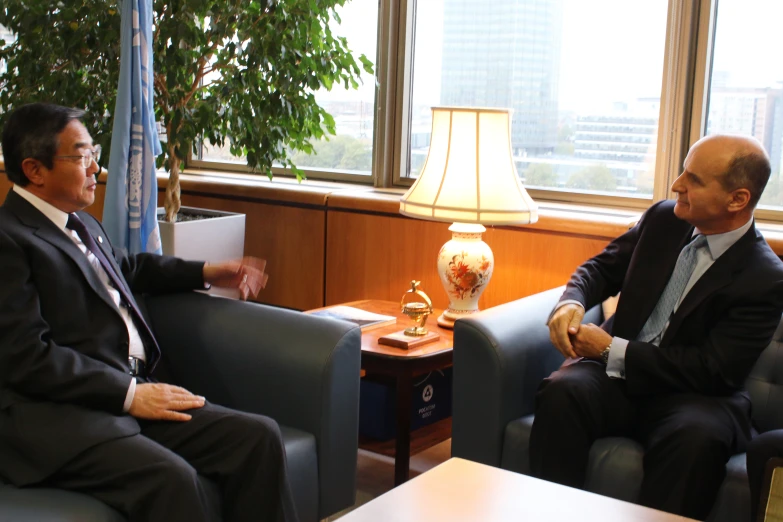 two men in suits are sitting in blue chairs and talking to each other