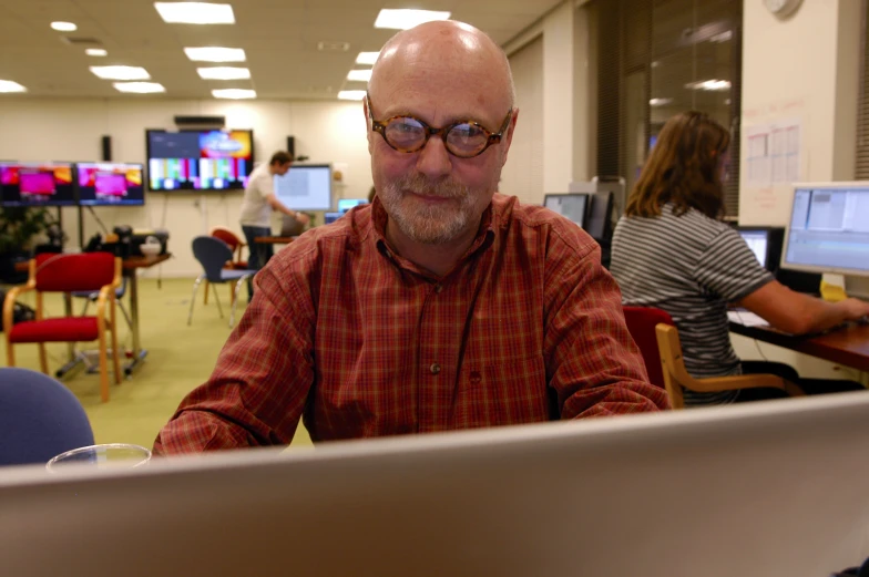 man with eyeglasses sitting at computer with people working in the background