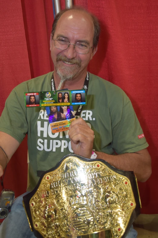 man in green shirt sitting next to red curtain holding up a large gold and silver belt