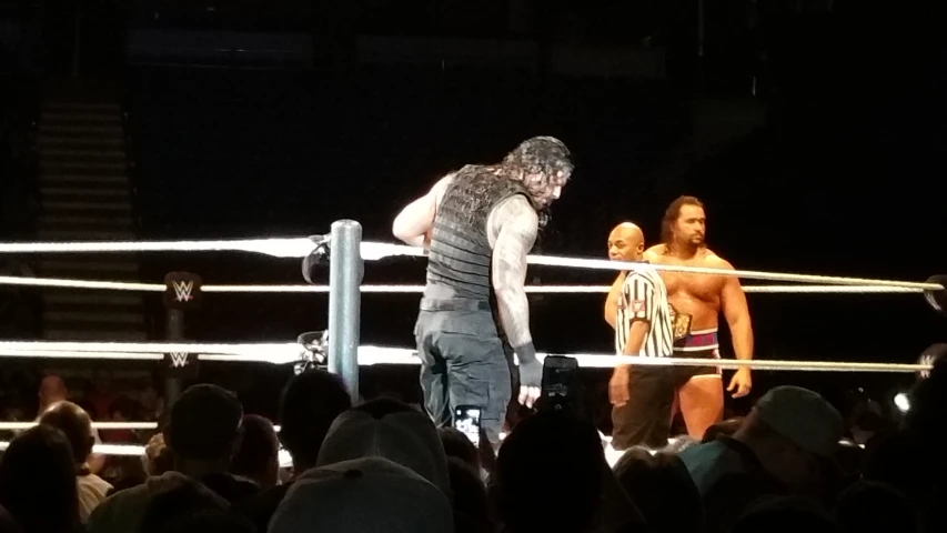 two men with short hair stand on one edge and look down as others watch from the other end of a wrestling ring