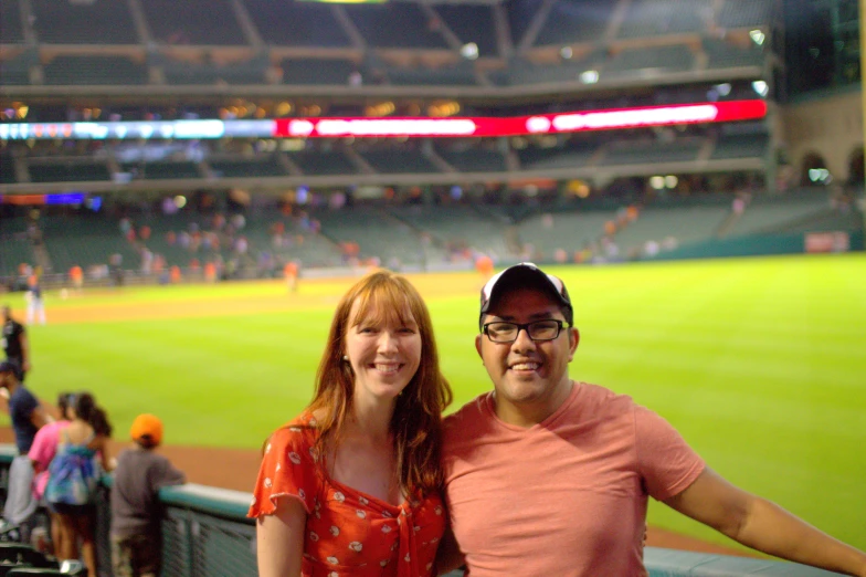 a couple poses for a picture in front of the empty baseball field