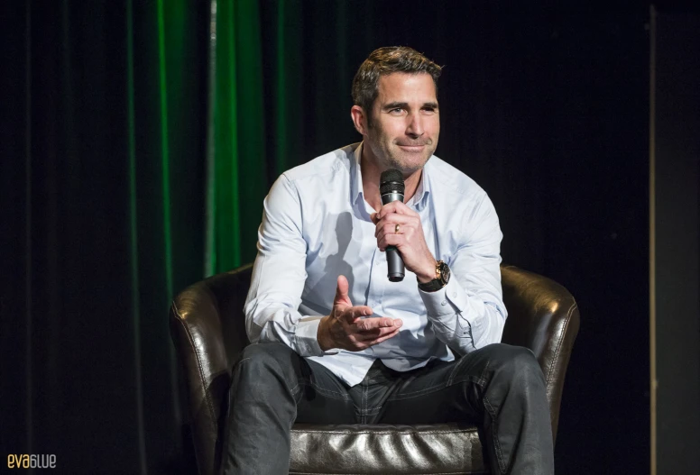 a man in a white shirt and black pants sits on a chair talking into a microphone