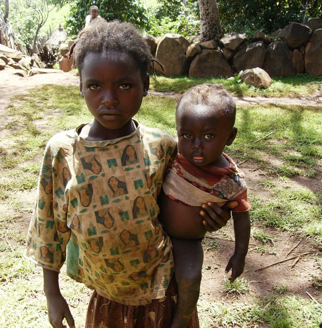 two little girls standing in the grass with one holding a baby