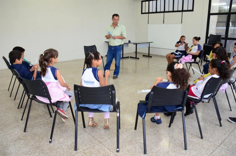 several children sitting in chairs in a classroom with a man standing by a table