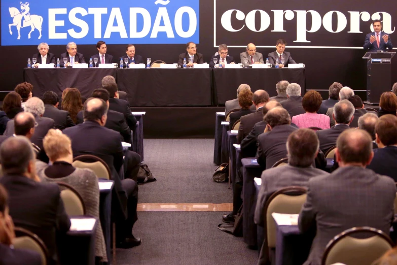 a group of people in chairs sitting at tables with microphones and banner in front of them