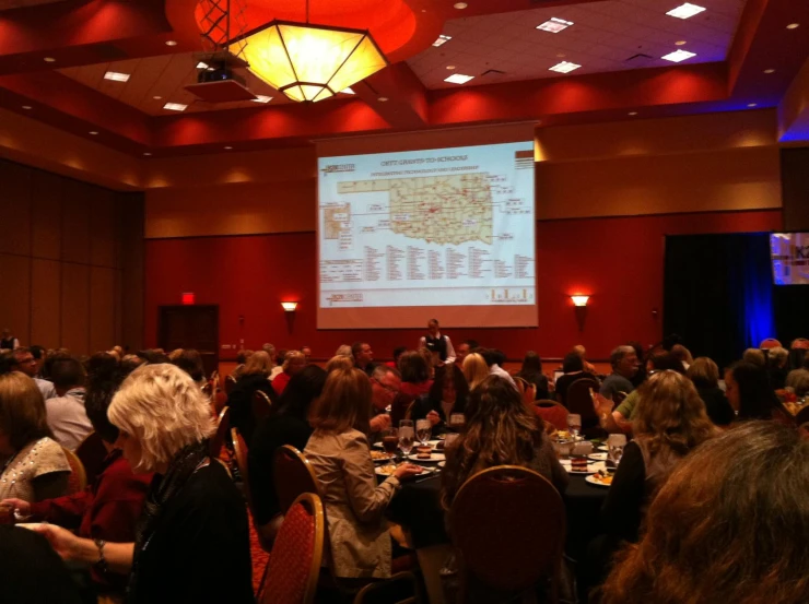 a large group of people sitting at tables in front of a projection screen