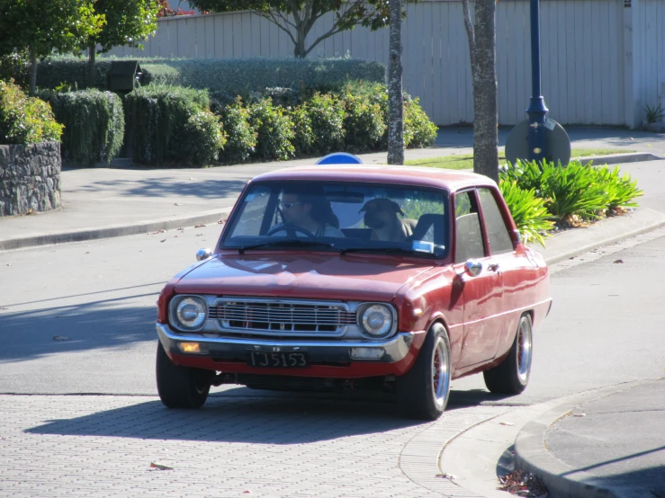 a red car driving down a street past bushes