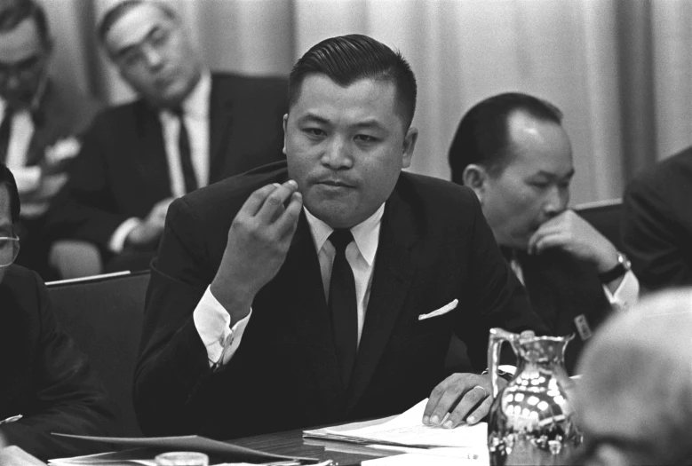 an asian man in a suit looks at soing while sitting at a desk