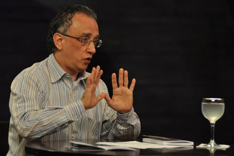 a man wearing glasses sitting at a table holding up hands