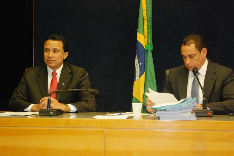 two men at desks in front of flags