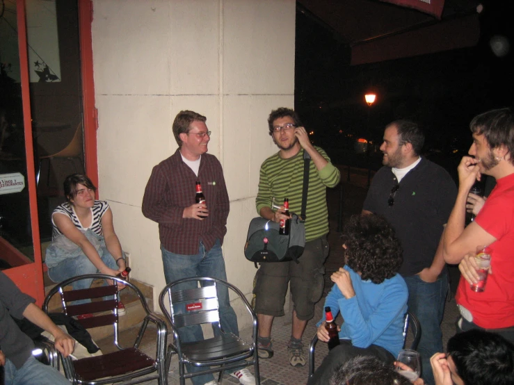 a group of men stand on a sidewalk next to a wall, while others sit around the bench holding onto their bottles