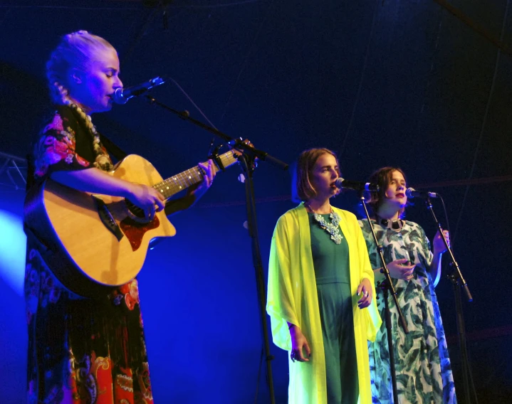 three women with guitars in front of microphones and on stage