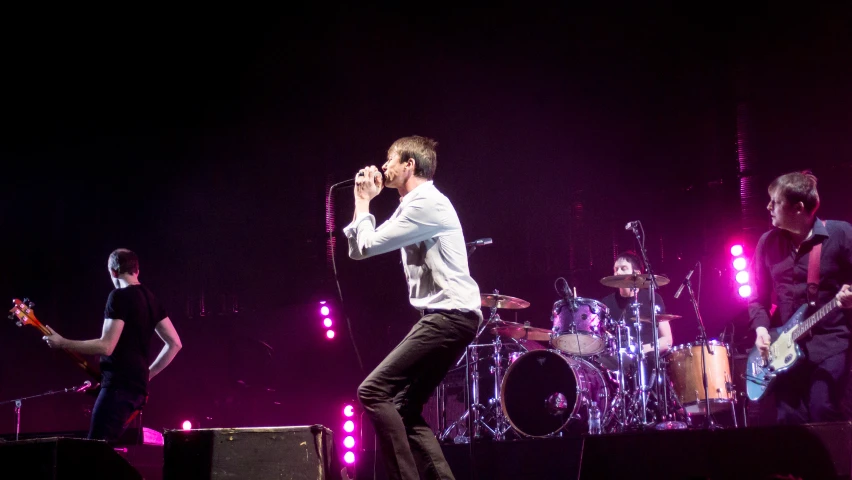 man in white jacket singing into microphone with band on stage