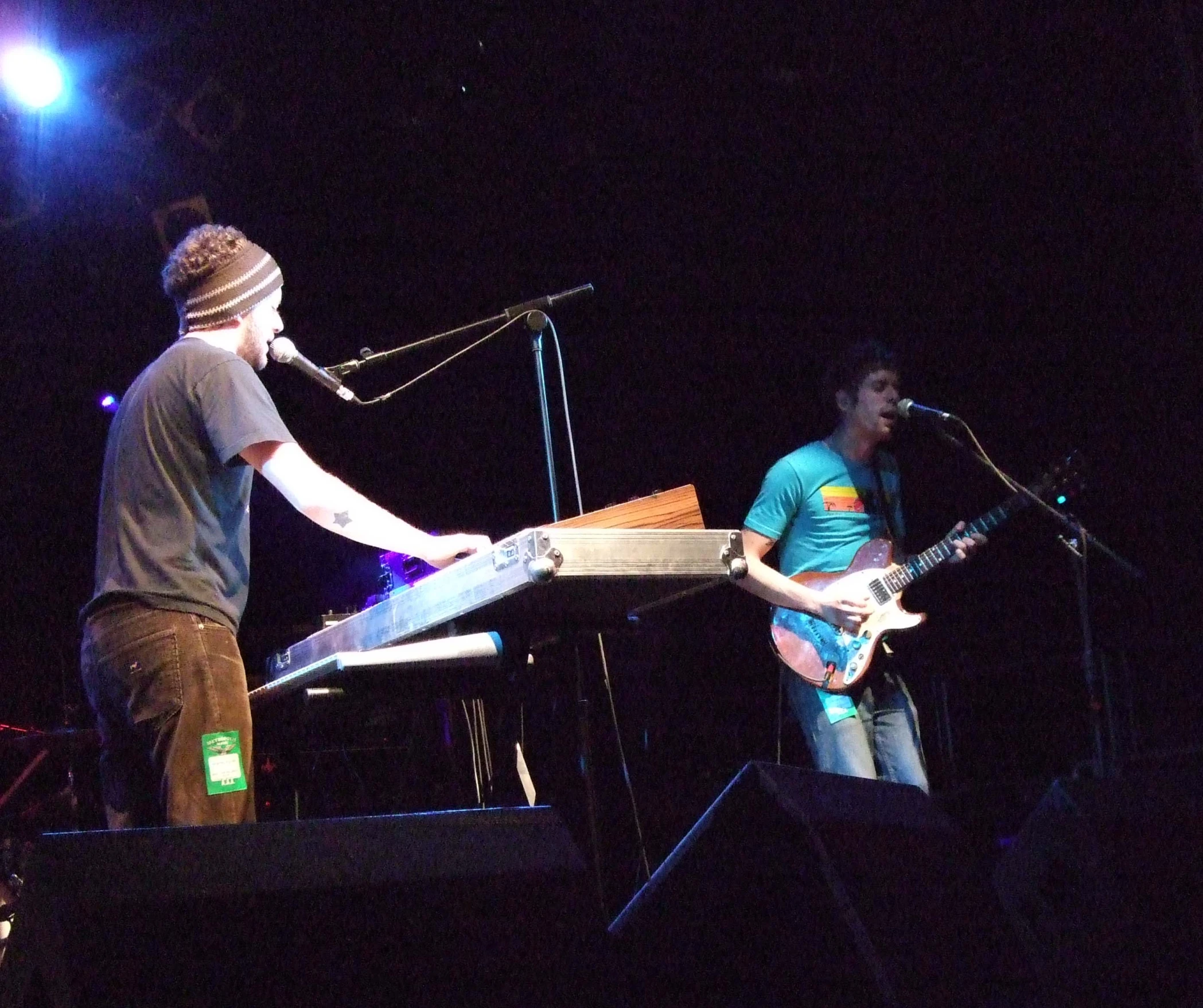 a man playing an electric guitar in front of a man with his arms folded out