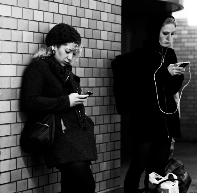 two women stand side by side against a brick wall, looking at their cell phones