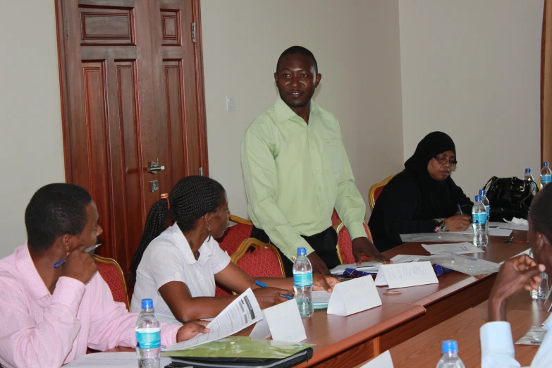 a man stands in the middle of a room with people seated at the table