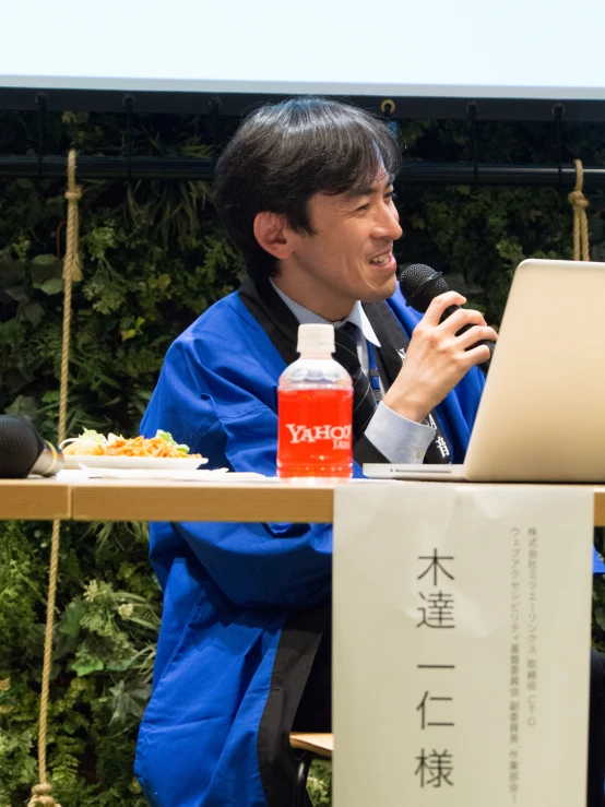 an asian man sitting in front of a laptop talking on a mic