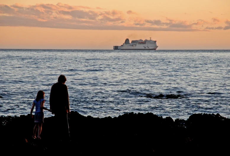 a man and a child standing near an ocean with a boat in the background