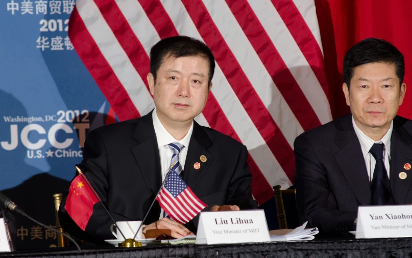 two asian businessmen sit side by side in front of american flags