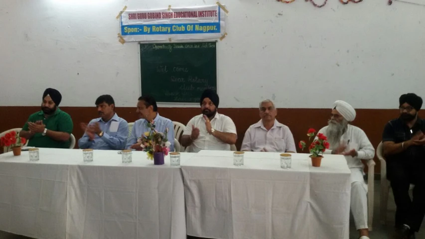 seven men sitting at a table with one person wearing a turban and the other man holding a flower