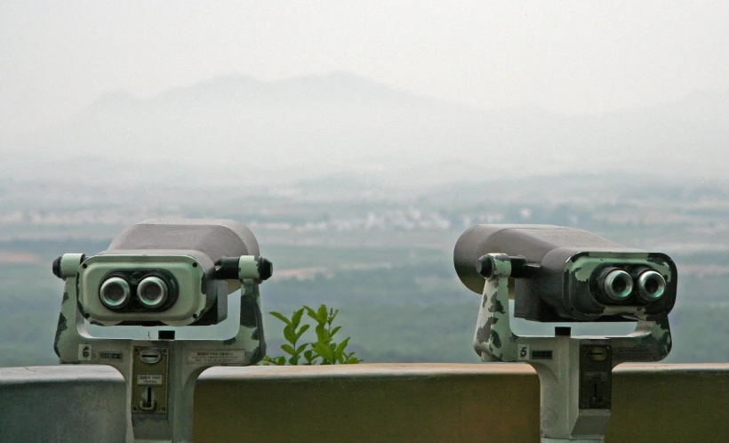 binoculars stand on the side of a wall with a view of an overcast hillside and a city