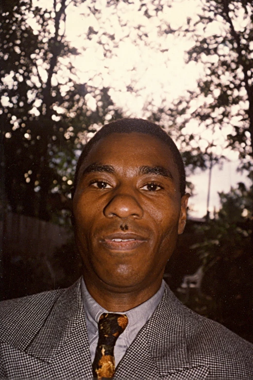 a black man in suit and tie with trees and building behind him