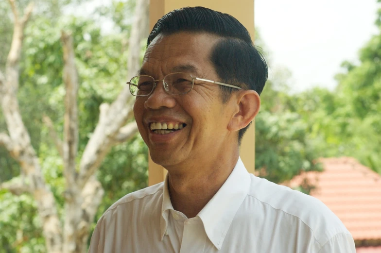 an asian man smiling outside with trees and sky in the background