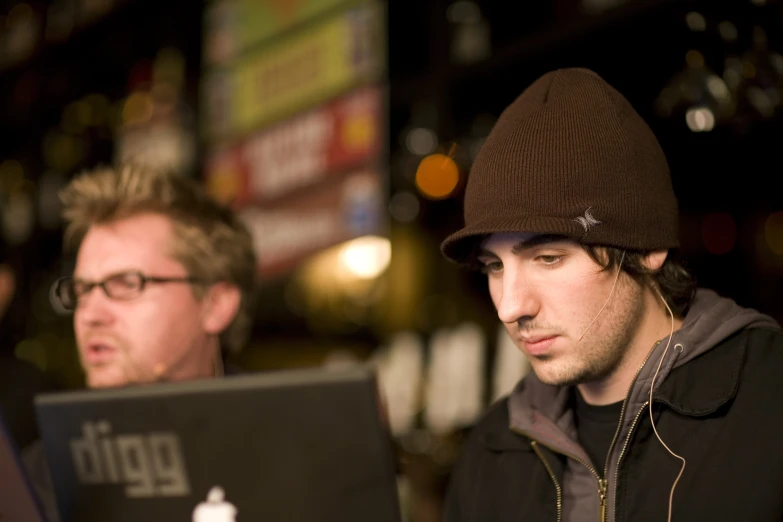 two men wearing winter jackets and beanies while using their laptops