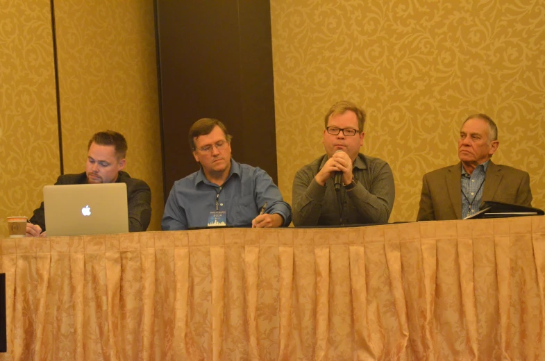 four men in business clothes sitting at a table with an apple laptop