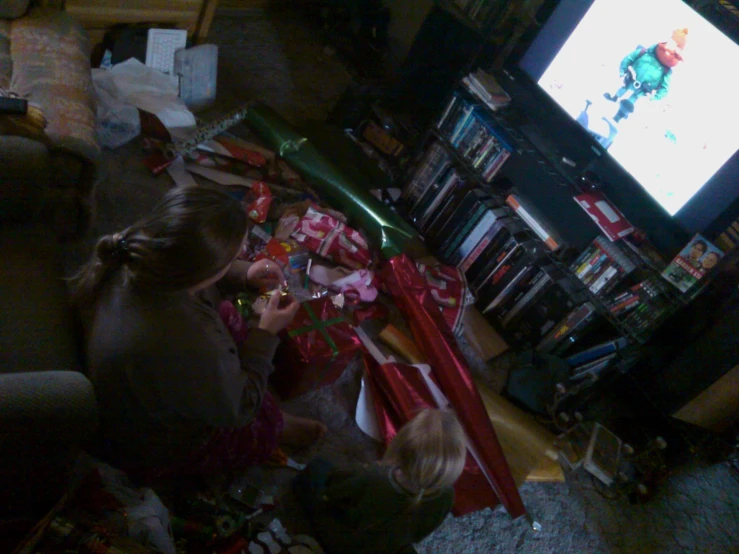 two children hold wrapping paper over christmas presents while looking at a television