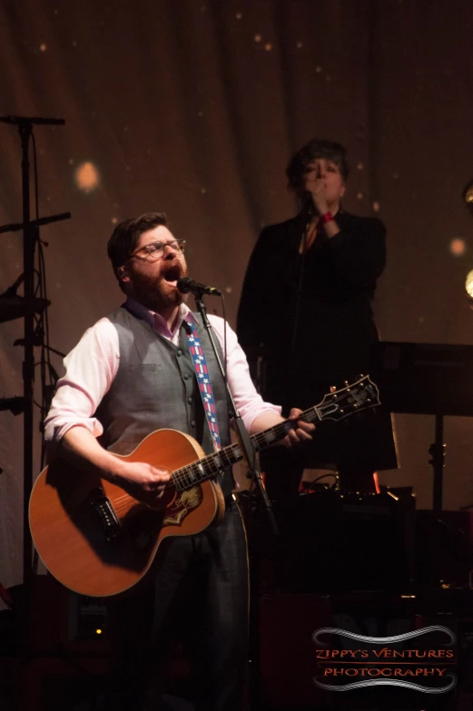 the man in grey vest sings while holding an acoustic guitar