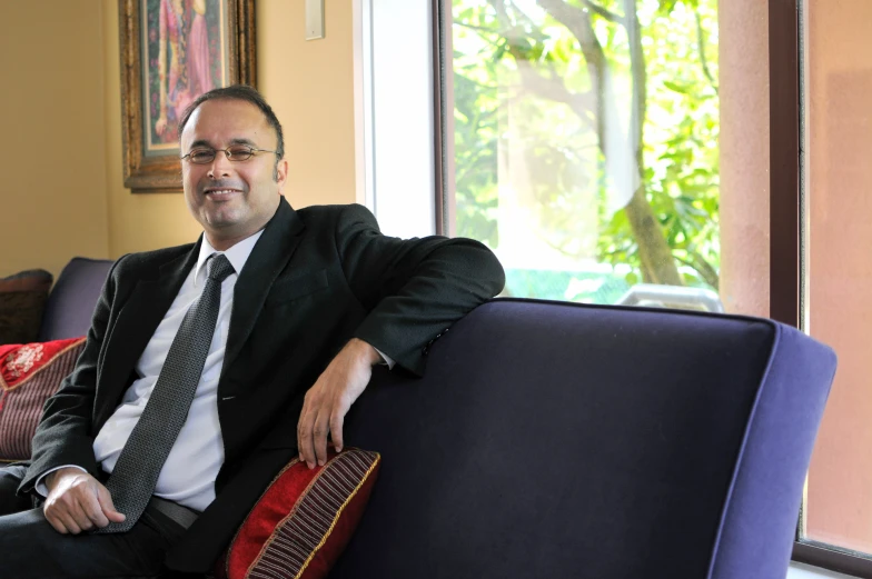 a man in a suit sitting on a purple chair with red and gold pillows