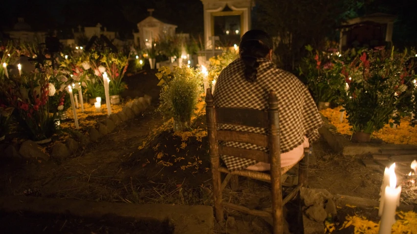 a woman sits outside a cemetery on a bench as the lighted candles glow