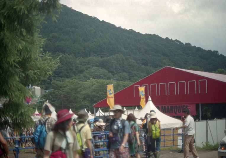 several people walking around a festival with tents and large trees in the background