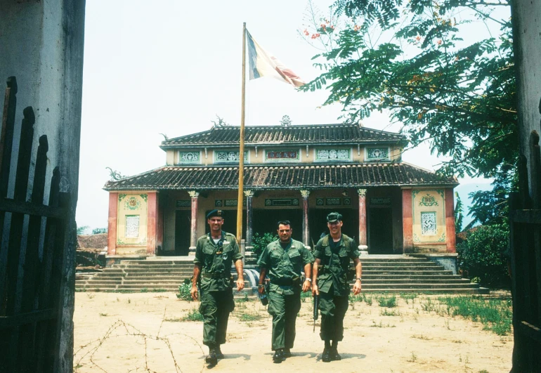 two men wearing fatigues are walking towards an old building