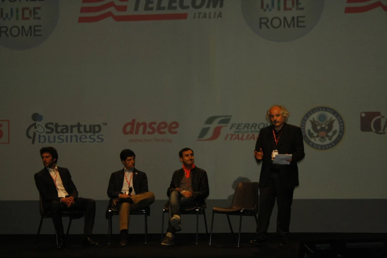 four men are in conversation on stage in front of a wall