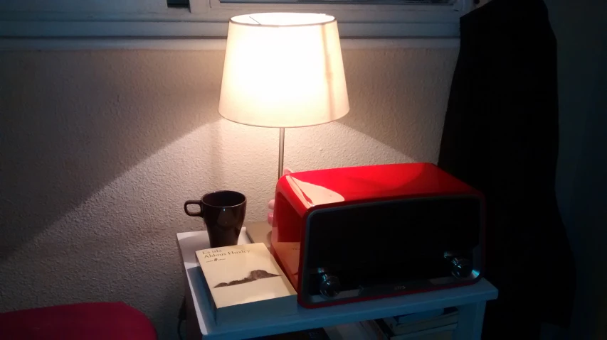 a white lamp, a coffee mug and a magazine sit on a small night stand