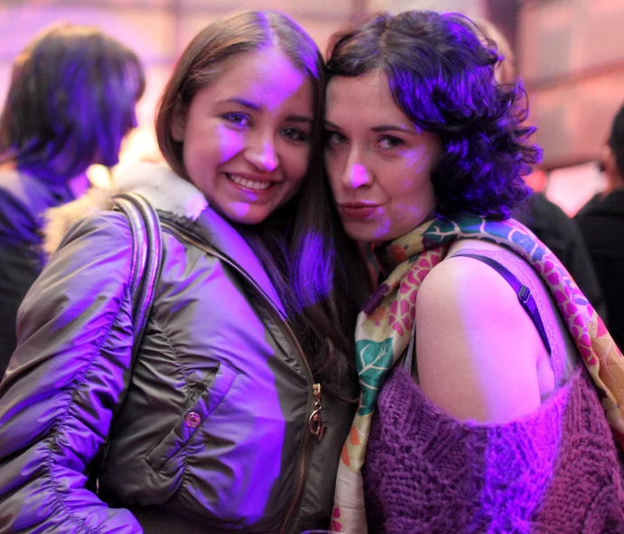 two women smile brightly at the camera, one holding her arms around the other