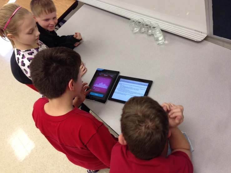 three young children sitting at a table playing on an electronic device