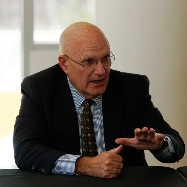 an older man with glasses and a suit, gesturing while sitting at a table