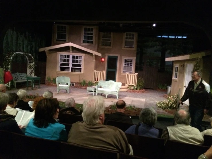 people sitting in front of small houses on stage with one man speaking