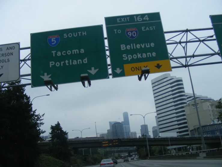 two highway signs on a highway overpass near trees and buildings