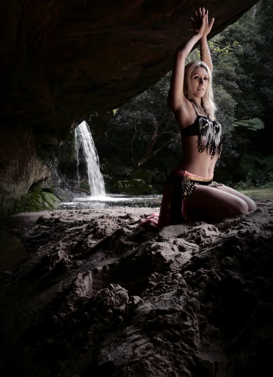 a young woman wearing a wet suit and hula skirt crouches under an unfurnished rock