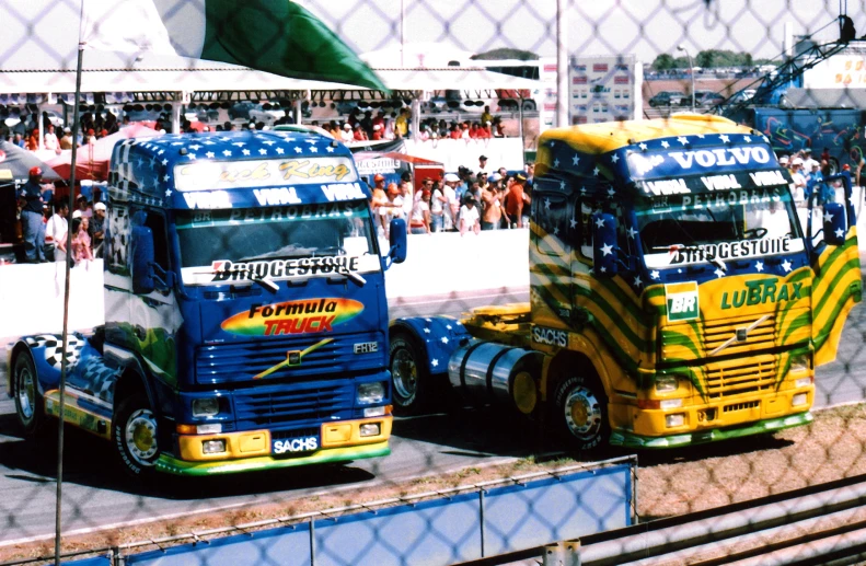 two big trucks driving down the road near a crowd