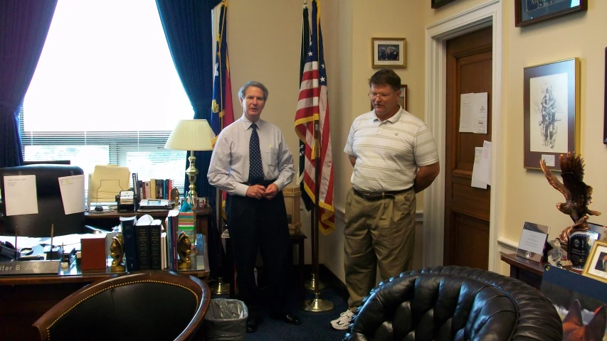 two men standing in a room next to two american flags